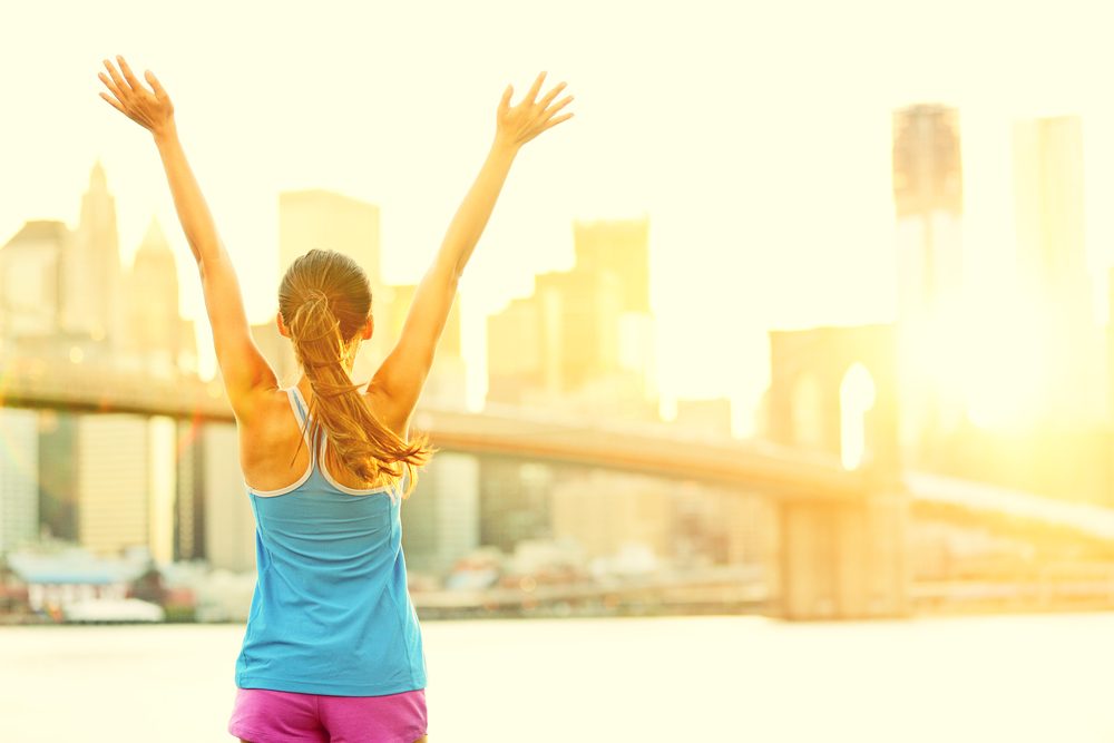 Woman in blue top with arms raised, facing a sunlit city skyline — hypnotherapy case study of a fitness fanatic who finally gave herself permission